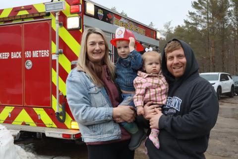 Young family with a child in each of the parents arms after visiting with Tiny Fire, standing in front of a fire truck
