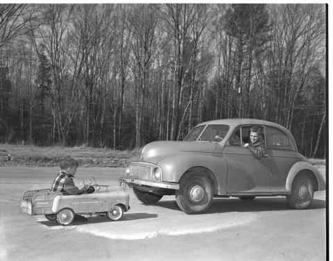 Classic car with person leaning out the window and looking at child sitting in toy car