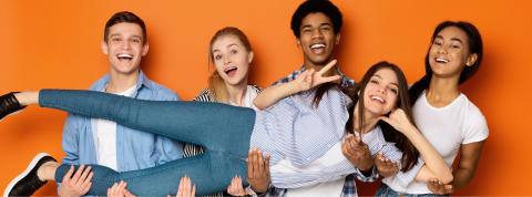 group of youth standing in front of a bright orange background. One of them is laying down and being held up by the others
