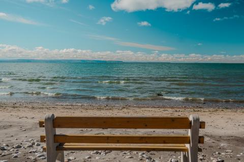 view from behind a public wooden bench looking out onto the beach with the lake slowly rolling waves into shore under a blue sky with light cloud cover