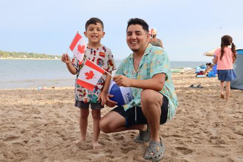 Father and son holding Canadian flags wearing brightly coloured clothing on the sand.