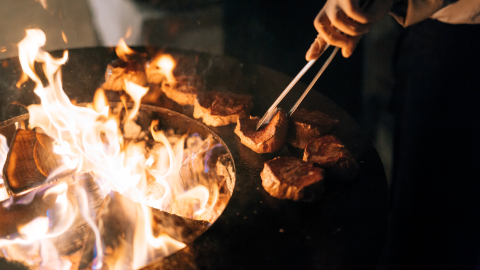 A person's hand holding tongs flipping pieces of meat on a fiery grill at night.