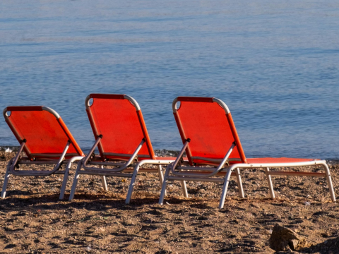Three empty red and white lounge chairs lined up on a sandy beach near the water's edge.