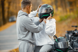 Man helping woman put on a helmet while she is sitting on one of the two ATVs parked on the side of the road